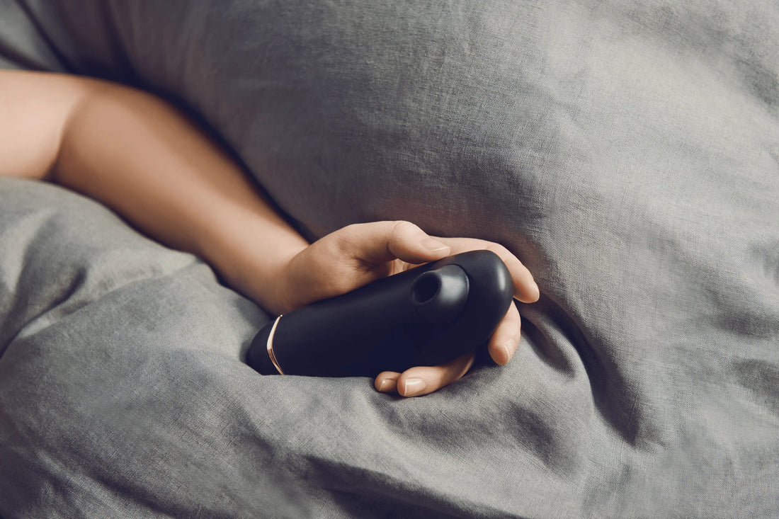 Hånd hviler roligt i sengetøj med sort Womanizer i grebet. Det ikoniske lufttryksstimulerende intimprodukt signalerer tryghed, nydelse og selvforkælelse i et afslappet, sanseligt øjeblik under dynen.