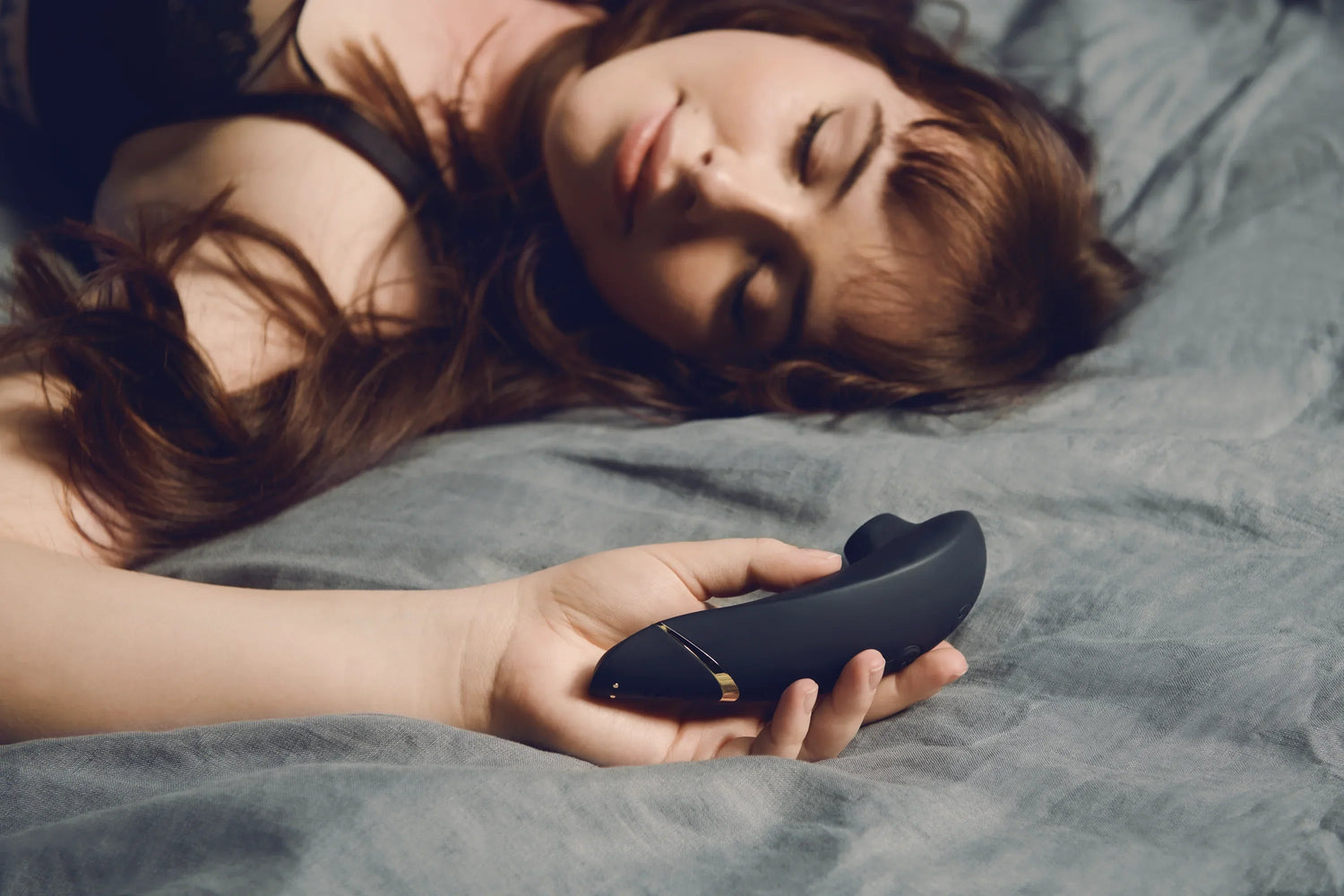 Kvinde ligger afslappet i sengen med en sort Womanizer lufttryksvibrator i hånden; stemningsfuldt billede til Womanizer Bundles kollektionen hos Nevola, der kombinerer vibratorer med glidecreme og pleje for fuldendt nydelse.