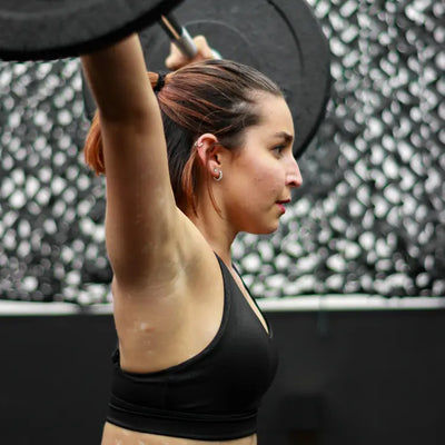 Kvinde i sort sports-bh løfter vægtstang over hovedet under styrketræning i fitnesscenter, set fra siden, med fokus på teknik og koncentration.