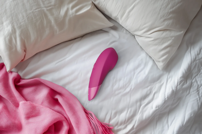 Womanizer Enhance placeret på en hvid seng med et rosa tæppe, som fremhæver produktets elegante form. Billedet viser den bløde, ergonomiske vibrator i en afslappet hverdagssituation, klar til rolig nydelse.