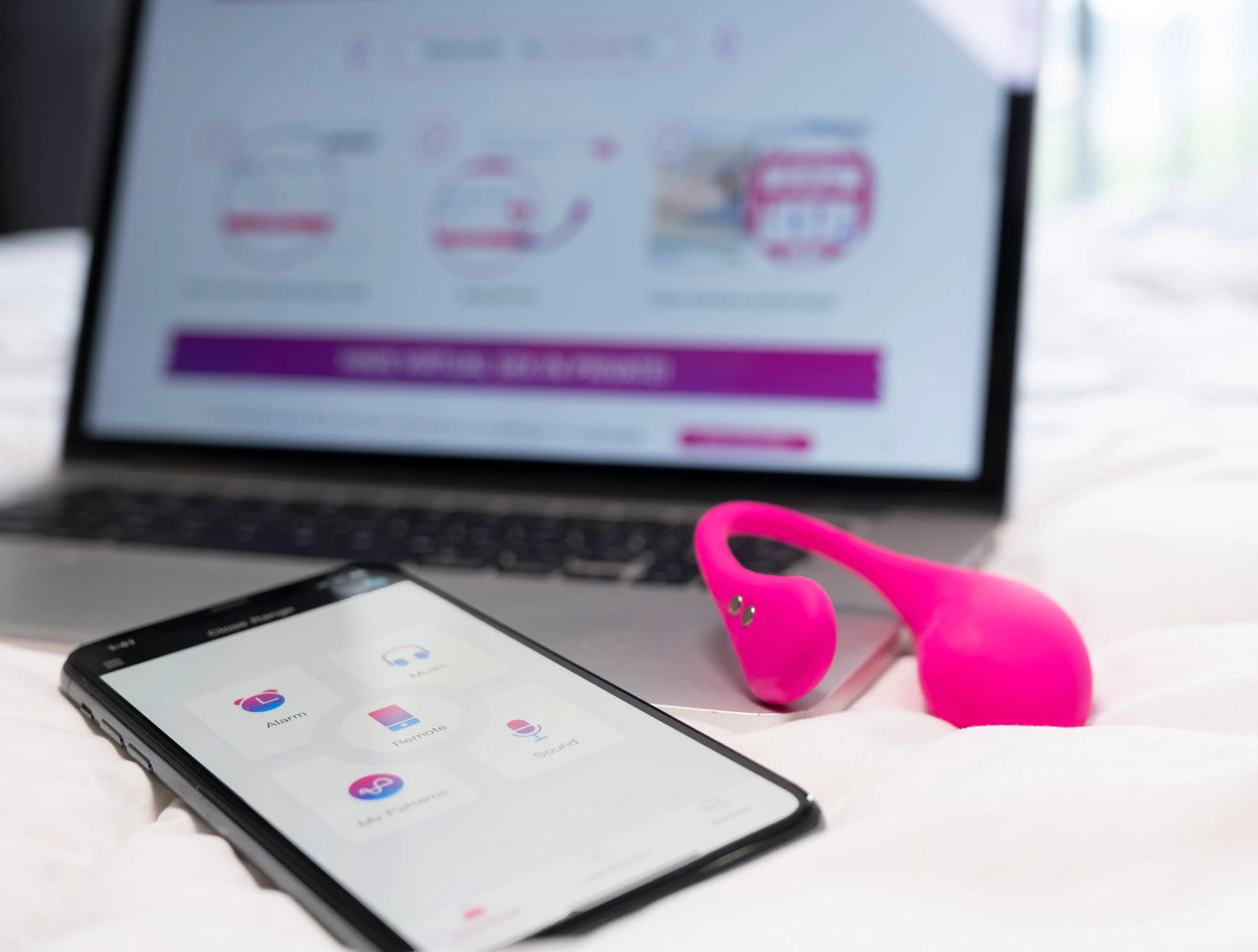 En stiliseret opsætning på en seng med den pinke Lush 3-vibrator i forgrunden, en smartphone til venstre med appens ikoner for funktionaliteter som Alarm, Remote og Music, og en MacBook til højre med Lovense-appens "Close Range"-side åben i baggrunden. Sort undertøj er synligt i hjørnet for at understrege det intime, parrettede design.