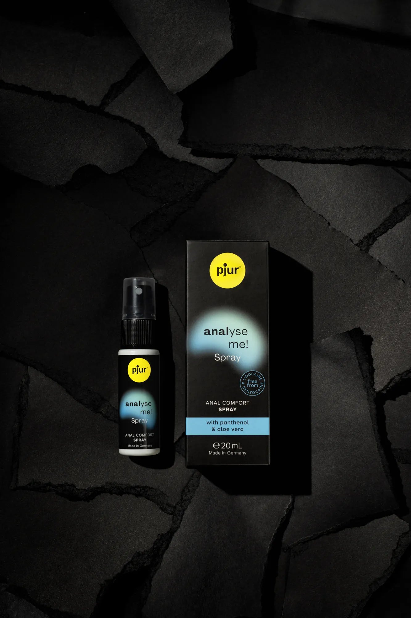 Et elegant produktbillede af pjur Analyse Me! Spray, der viser en sort pumpeflaske (20 ml) med blå etiket og en matchende sort boks. Etiketten markerer "ANAL COMFORT SPRAY with Panthenol & Aloe Vera" og "Made in Germany". Boksen har blå tekster som "Relaxation spray for anal sex" og "reduces sensitivity". Billedet er sat op på en sort, revnet overflade med subtile blå nuancer for at formidle afslapning og komfort under intim brug.