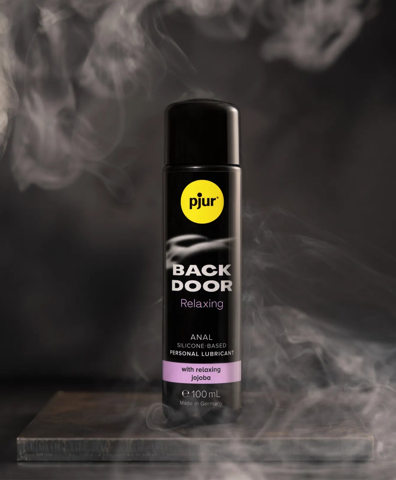 Flaske med Pjur Back Door Relaxing anal glidecreme placeret på mørk overflade med damp omkring; produktbillede fra Pjur kollektionen hos Nevola, der fremhæver silkeblød, silikonebaseret komfort med afslappende jojoba.