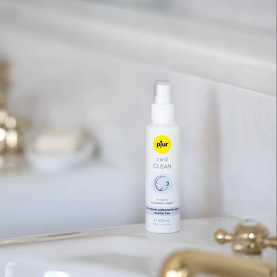 Pjur Med Clean rengøringsspray placeret elegant på badeværelse med gyldne detaljer. Lifestyle-billede der viser diskret og stilfuld pleje af intimprodukter i en rolig, naturlig hverdagsscene.