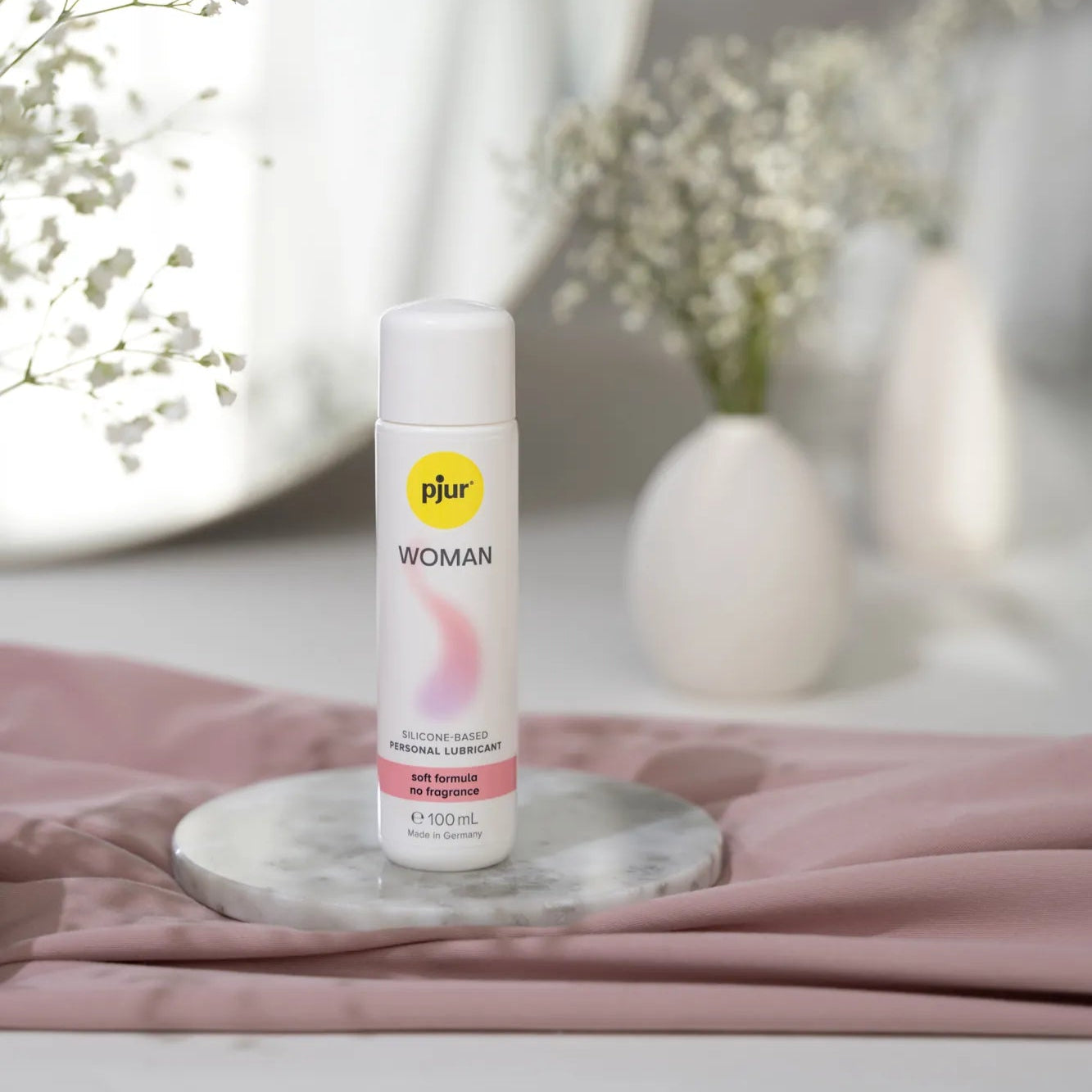 Pjur Woman silikonebaseret glidecreme i 100 ml flaske fotograferet i elegant miljø med bløde tekstiler og blomster. Plejende, parfumefri formel udviklet til sensitiv hud, giver langvarig silkeglat nydelse.
