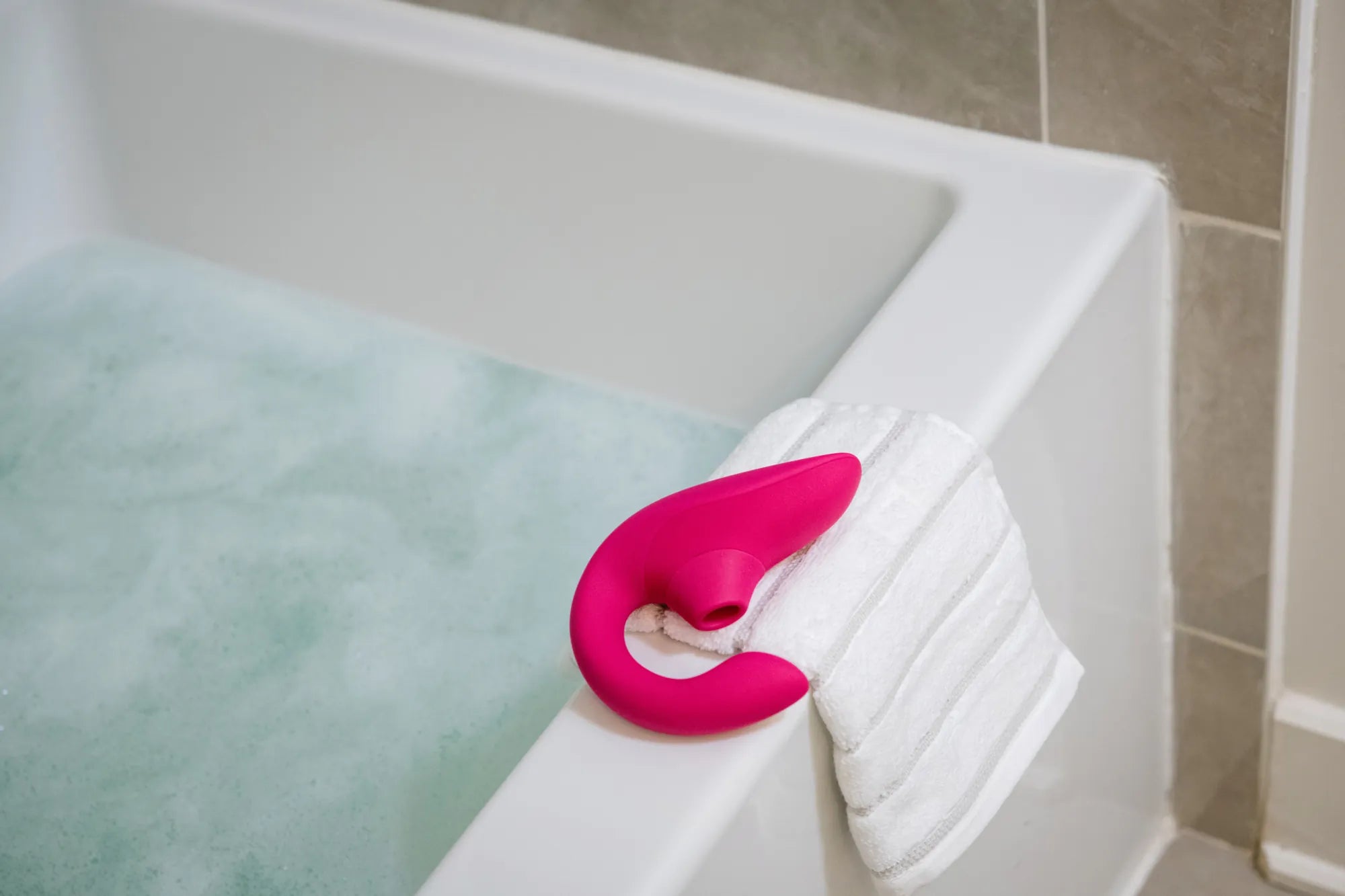 Womanizer Blend sexlegetøj i pink ligger på kanten af et badekar sammen med et håndklæde. Billedet viser lufttryksvibratorens vandtætte funktion og afslappende spa-inspireret brugssituation.