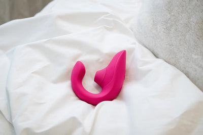 Womanizer Blend klitorisstimulator i pink ligger på hvide lagner i et roligt soveværelsesmiljø. Billedet fremhæver sexlegetøjets elegante buede design og lufttryksfunktion til sanselig stimulering og diskret selvforkælelse.
