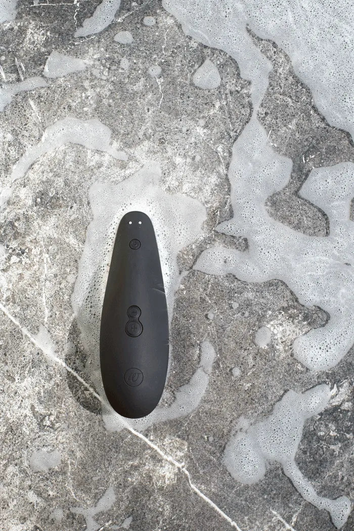 Womanizer Classic 2 lufttryksvibrator i sort liggende på stenet overflade med vand og skum. Eksklusivt sexlegetøj til kvinder, kombinerer diskret æstetik, Pleasure Air-teknologi og vandtæt kvalitet.