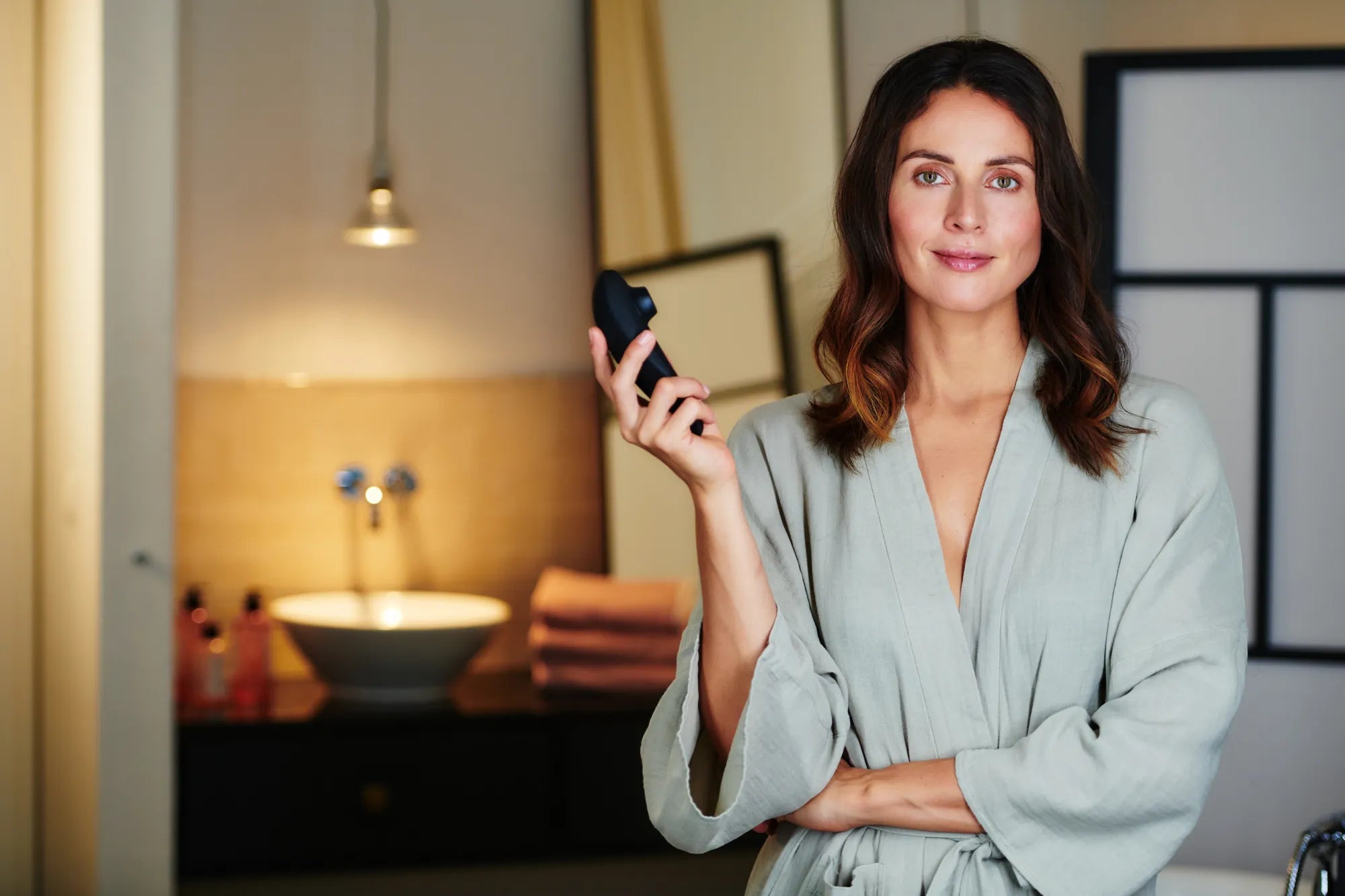Kvinde iført lysegrå morgenkåbe står roligt i badeværelset og holder en Womanizer Classic 2 i hånden; stemningsfuldt billede, der formidler velvære, selvforkælelse og diskret luksus fra Womanizer kollektionen hos Nevola.