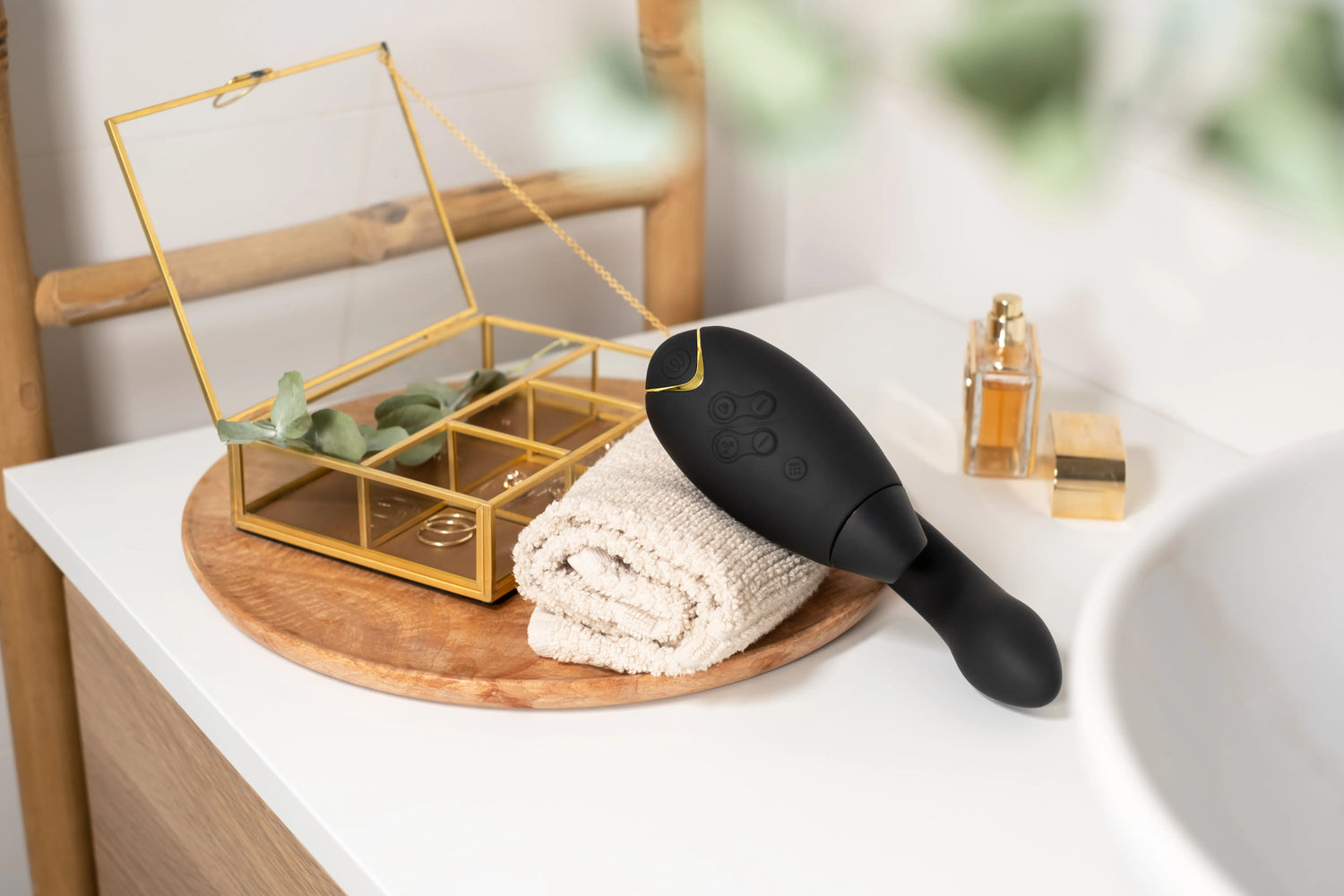 Womanizer Duo 2 i sort placeret på badeværelsesbord ved et blødt håndklæde og gyldent smykkeskrin. Eksklusivt sexlegetøj, der kombinerer stilfuldt design og funktionel dobbeltstimulering i hverdagen.