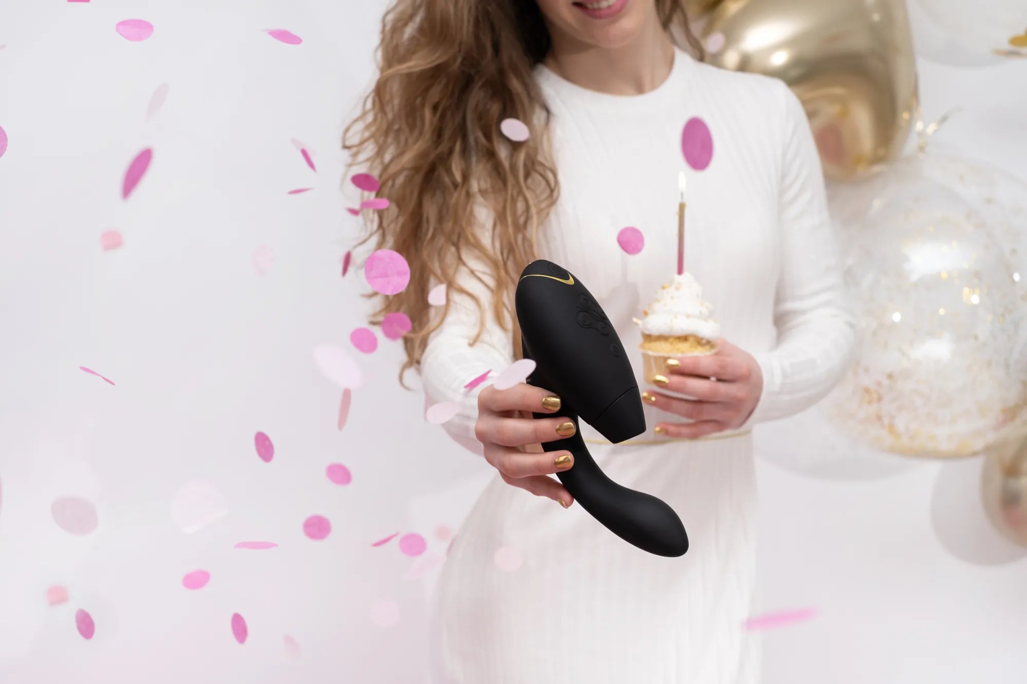 Kvinde i hvid kjole holder Womanizer Duo 2 sammen med en cupcake, omgivet af guld og pink konfetti. Premium sexlegetøj præsenteret i et sanseligt og legende univers.