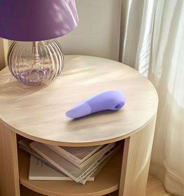 Womanizer Enhance i farven lilac placeret elegant på et natbord i soveværelse – diskret, stilfuldt og klar til intim nydelse, når du ønsker et øjeblik af selvforkælelse.