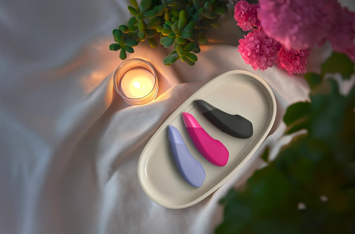 Tre Womanizer Enhance modeller i lilla, pink og sort placeret på et fad omgivet af blomster og stearinlys; stemningsfuldt billede, der viser elegante lufttryksvibratorer fra Womanizer kollektionen hos Nevola i et roligt, sanseligt miljø.