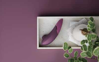 Sort Womanizer Next placeret på marmor fad ved siden af duftlys og grønne planter – eksklusivt sexlegetøj, der kombinerer design, teknologi og sensuel forkælelse.