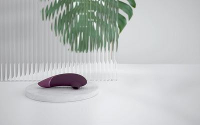 Womanizer Next i mørklilla farve placeret i dekorativ bakke ved duftlys og grønne blade – sofistikeret sexlegetøj med fokus på design og velvære.