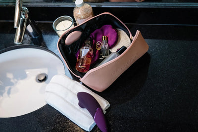 Åben kosmetiktaske med Womanizer Next, parfume og satinøjenmaske – et stilrent billede, der udstråler feminin luksus og personlig velvære.