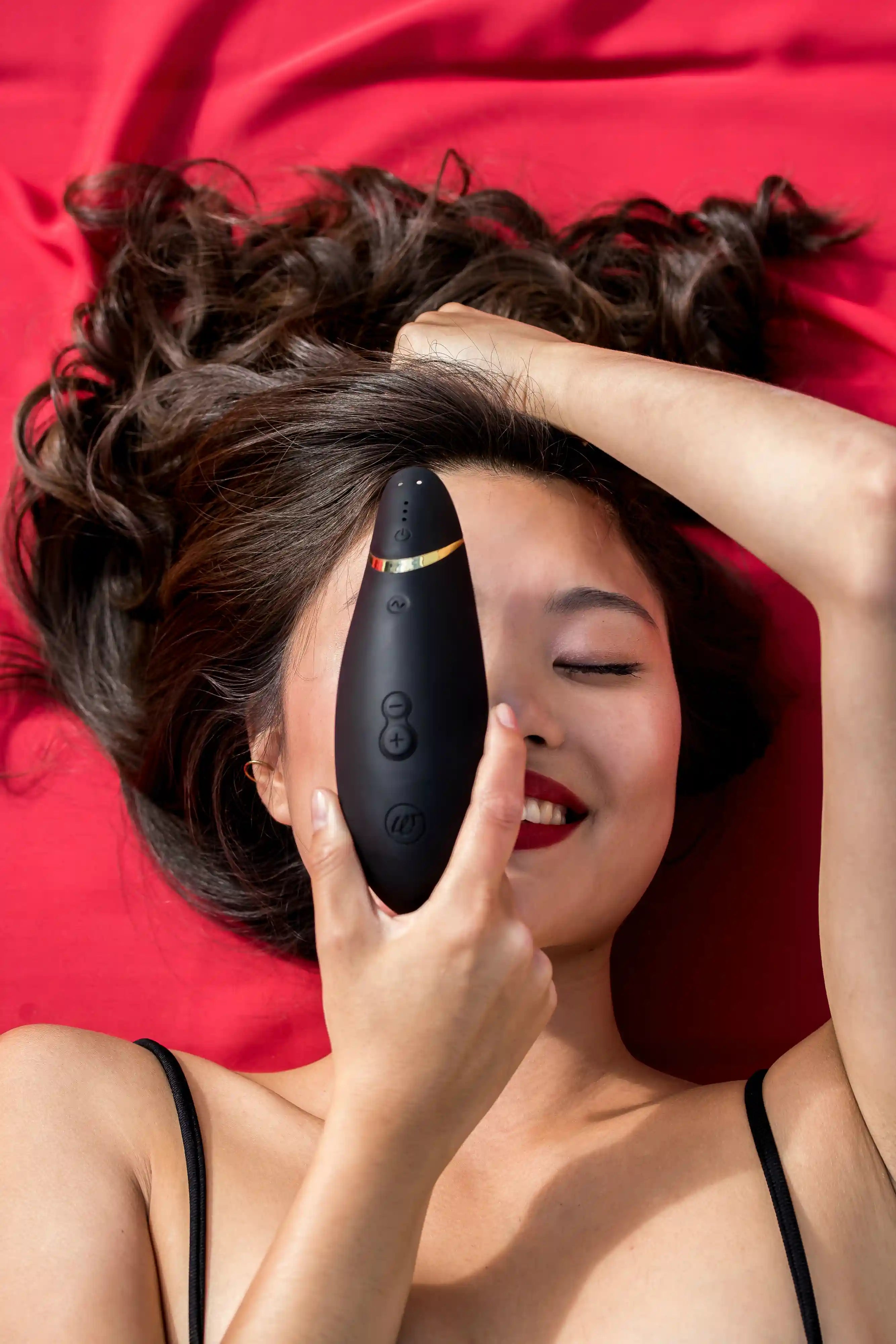 Kvinde ligger afslappet på rød baggrund med Womanizer Premium 2 i sort og gyldne detaljer. Sexlegetøjet stimulerer med lufttryk og vibrationer for sensuel, intens og stilfuld nydelse.