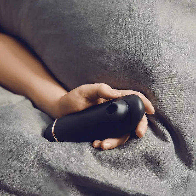 Sanseligt produktfoto af Womanizer Premium 2 i elegant sort design, holdt i en kvindes hånd under bløde dyner. Intimprodukt med lufttryksstimulering, der giver diskret, rolig og intens nydelse.