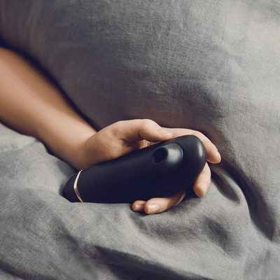 Stemningsfuldt billede af Womanizer Premium 2 sort, placeret i en kvindes hånd under sengetæppe. Lufttryksvibrator til kvinder, skabt til sensuel wellness og naturlig selvforkælelse i intime øjeblikke.