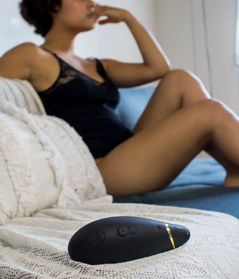 Womanizer Premium Black placeret på et badeværelse i en intim wellness-kontekst, mens en kvinde i baggrunden gør sig klar. Et diskret og luksuriøst sexlegetøj til kvinder, skabt til rolig nydelse og hverdagsforkælelse.