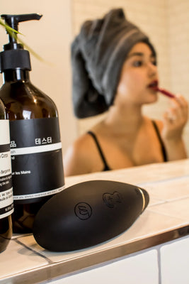 Womanizer Premium Black ligger i forgrunden på en seng, mens en kvinde i sort lingeri ses i baggrunden. Et elegant sexlegetøj til kvinder, der signalerer ro, sanselighed og eksklusiv selvforkælelse.